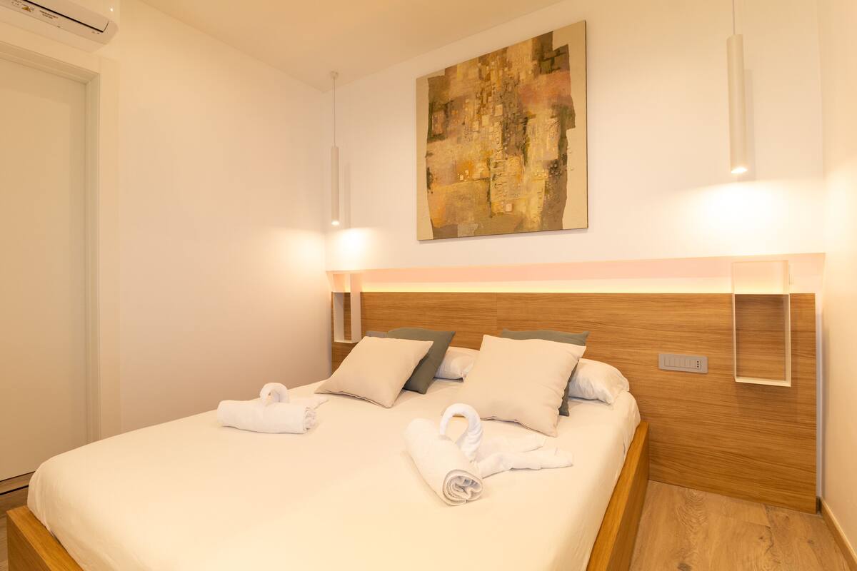 A hotel room with a neatly made bed, a headboard with built-in lighting, a painting on the wall, and a bedside table with a lamp.