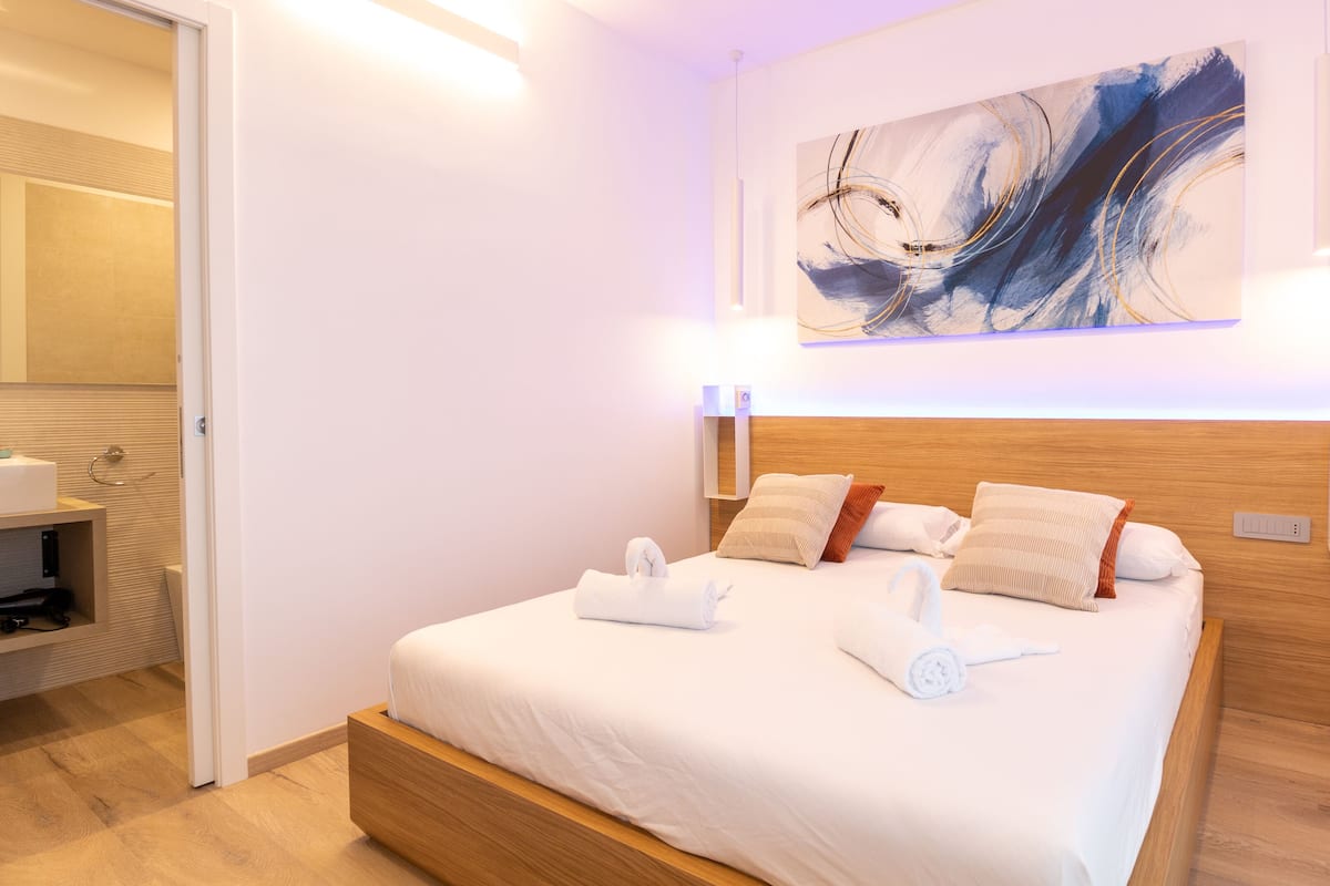 A modern hotel room with a large bed, wooden headboard, and abstract art on the wall.