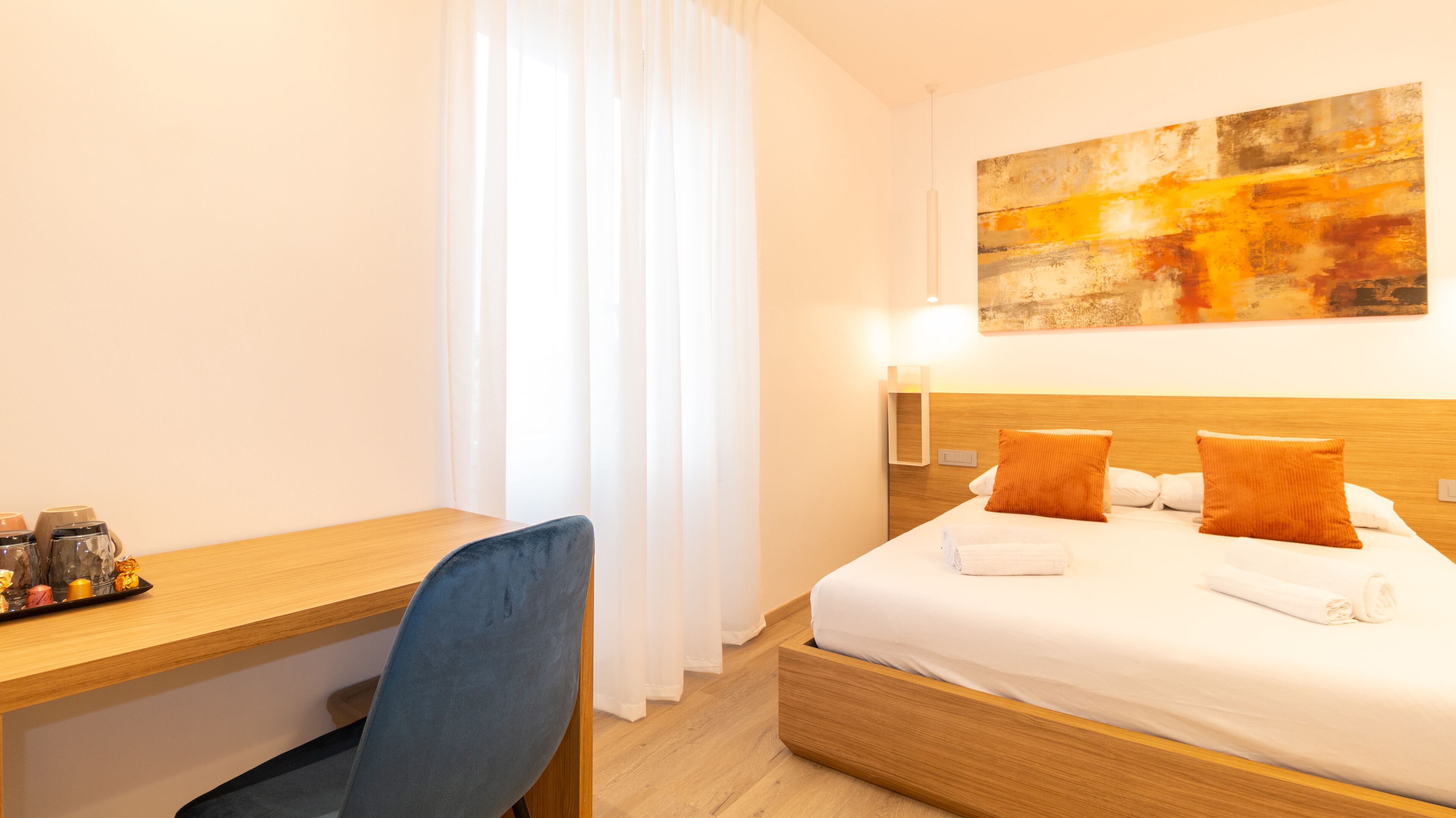 Une chambre d’hôtel avec un bureau en bois, une chaise bleue, un lit avec deux oreillers orange et un tableau au mur.