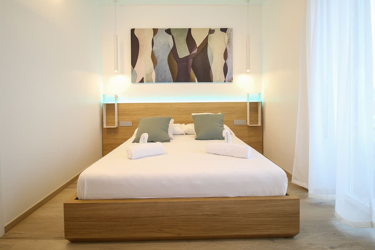 A modern hotel room with a wooden bed frame, white bedding, two pillows, and a wall-mounted abstract painting.