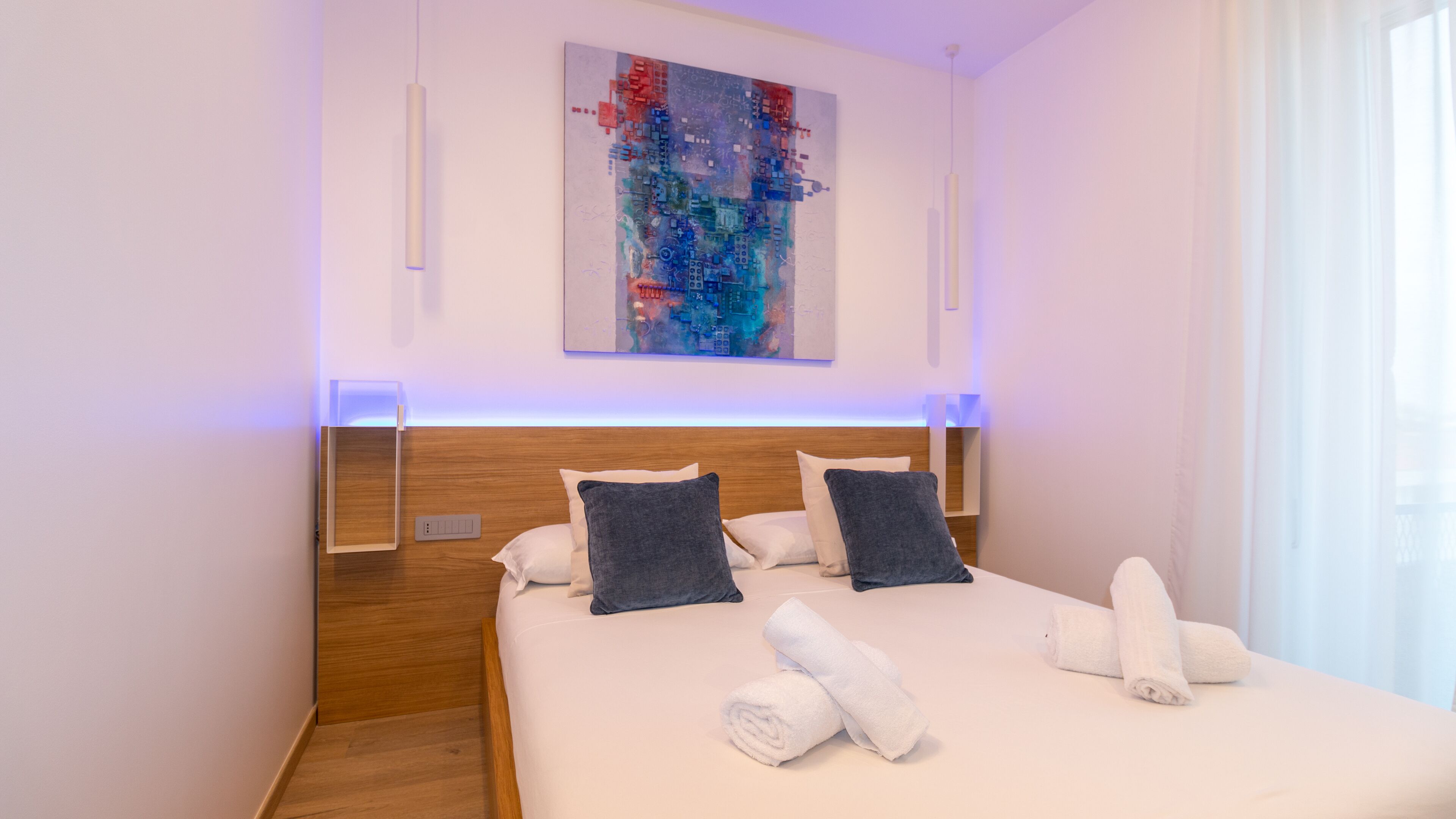 A modern hotel room with a wooden headboard, a large abstract painting on the wall, and neatly folded towels on the bed.