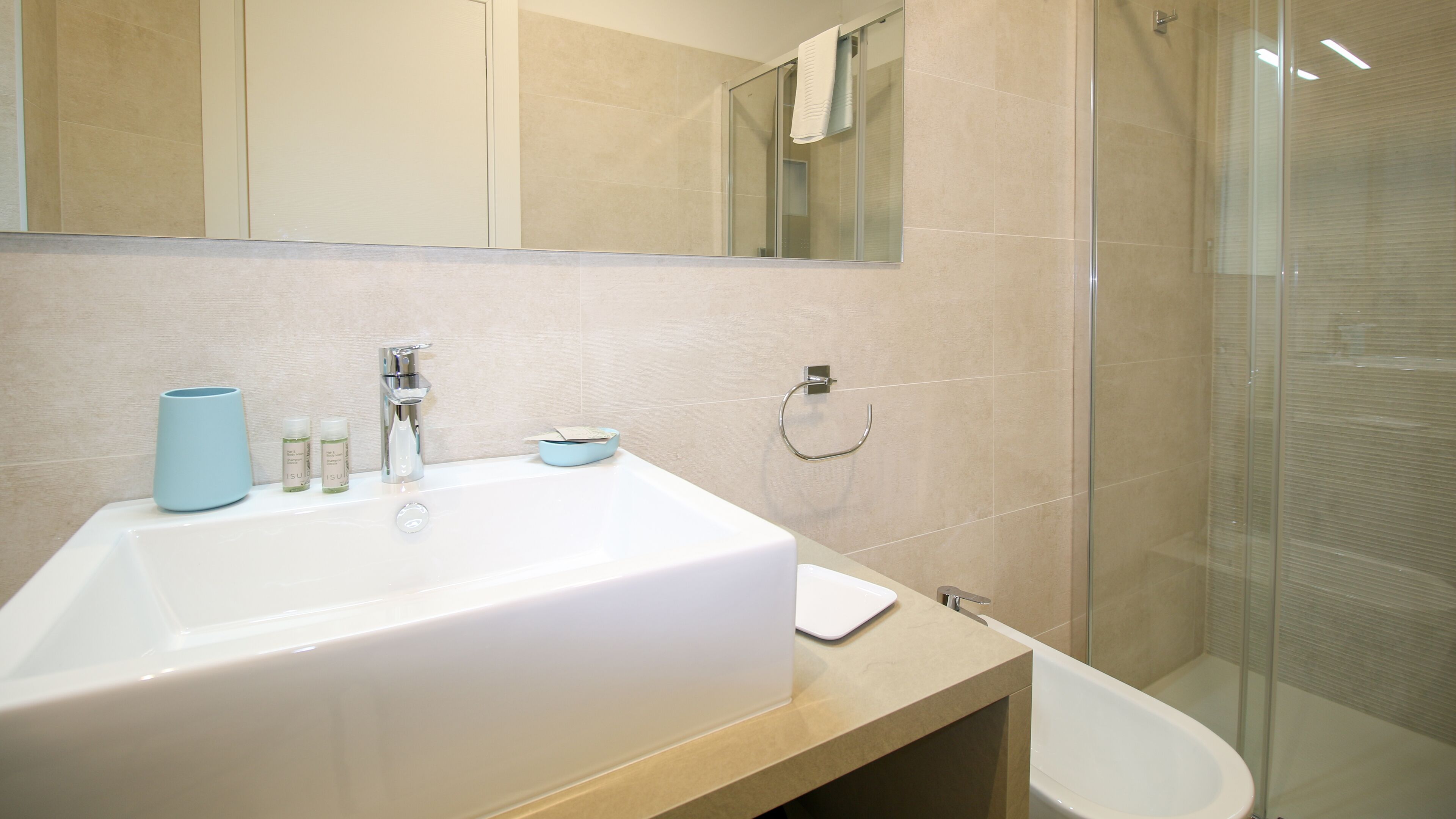 A modern bathroom with a large white sink, a glass shower enclosure, and a beige tiled wall.