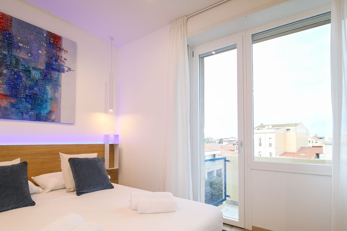 A hotel room with a large bed, a cityscape painting, a view of buildings through a balcony door, and a white curtain.