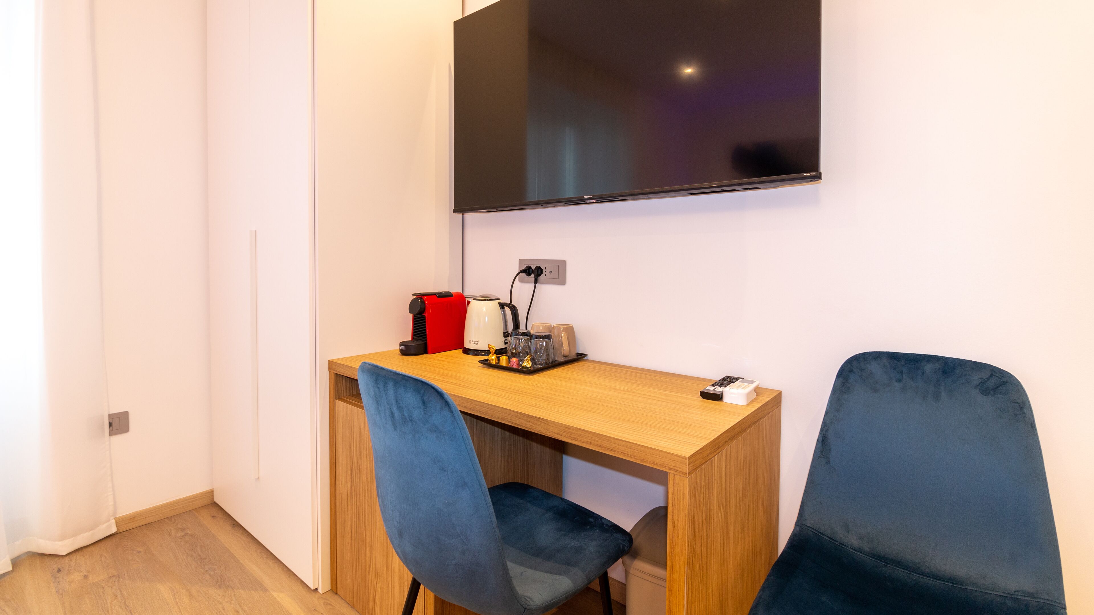 Une petite pièce avec un bureau en bois, deux chaises bleues, un téléviseur à écran plat fixé au mur, une chaise rouge, une machine à café et une bouilloire.