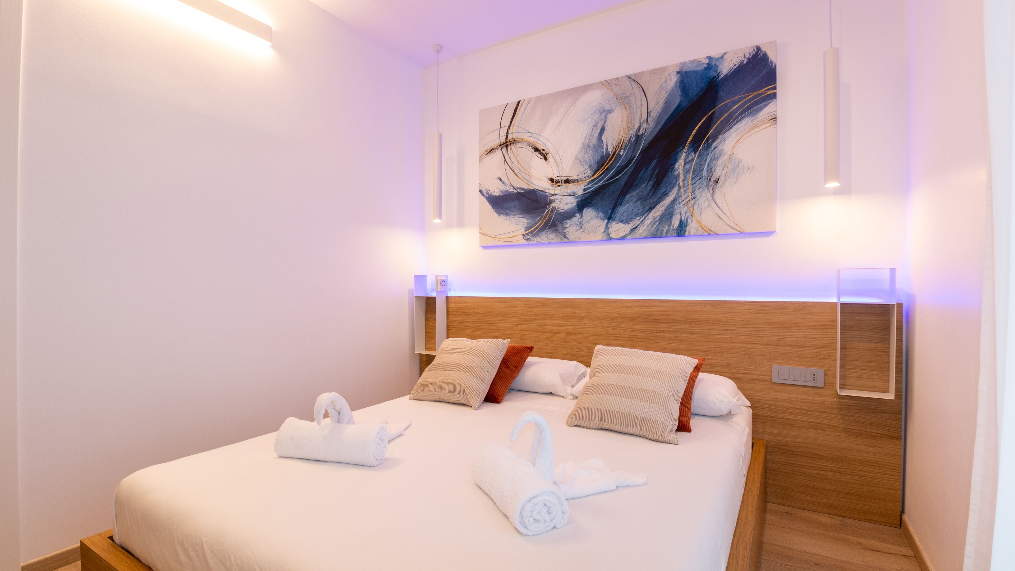 Una habitació d'hotel moderna amb un llit gran, roba de llit blanca i un panell de fusta al capçal. Hi ha un quadre sobre el llit i un aplíc a la paret.