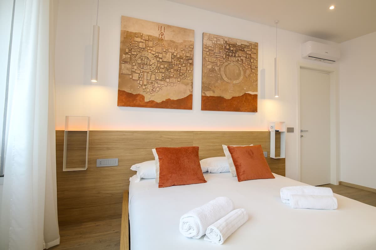 A hotel room with a large bed, two wall-mounted artworks, a wooden headboard, a bedside table with a lamp, and a door with an air conditioning unit.