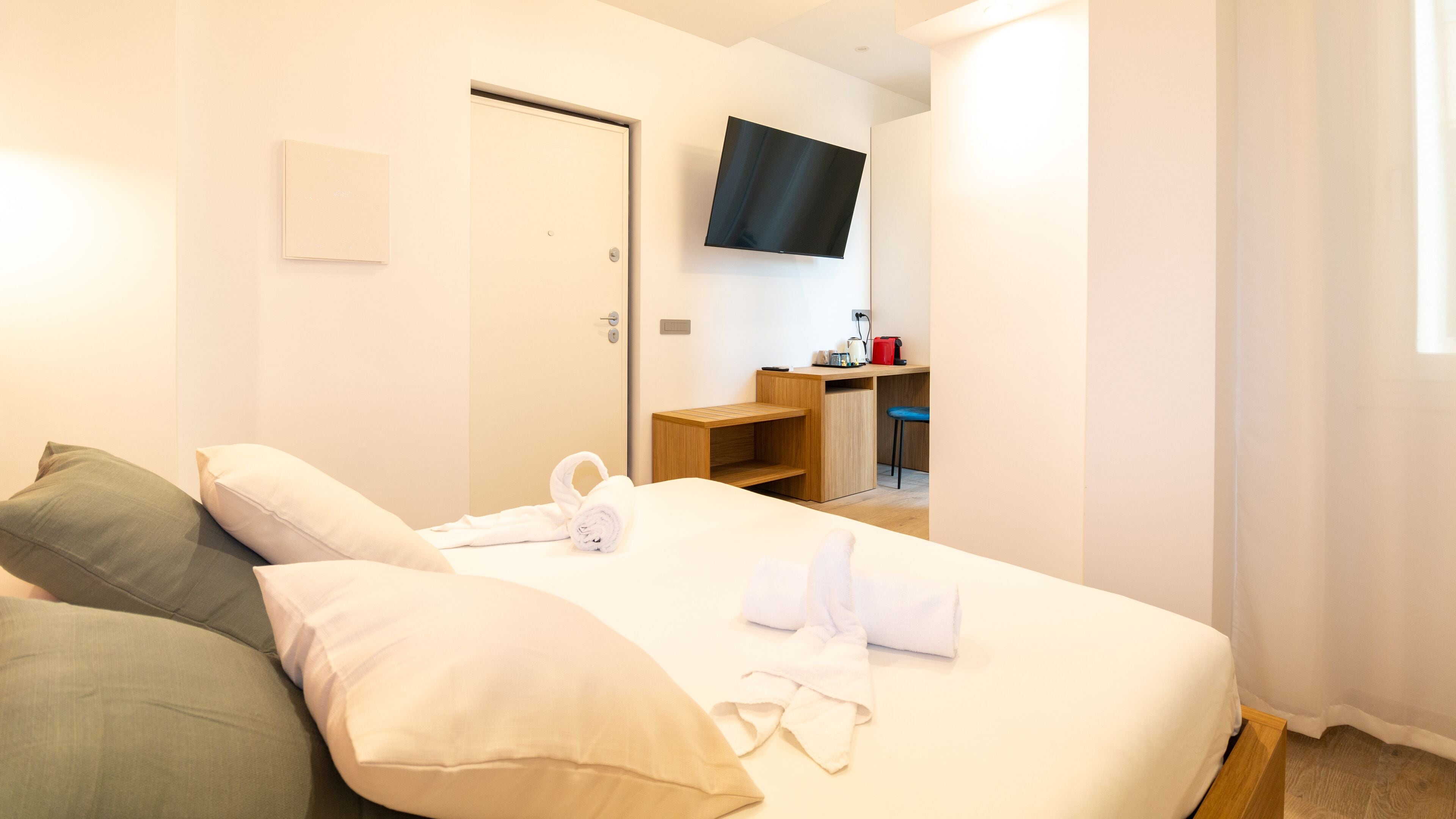 Une chambre d’hôtel comprenant un lit, des oreillers, un téléviseur fixé au mur, un bureau avec une chaise et une fenêtre avec des rideaux.