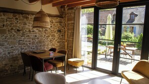 A stone-walled room with a table and chairs, a wicker lamp, and a view through glass doors to an outdoor patio area. - Oude Schuur Omgetoverd tot Moderne Gîte (montchevrier)