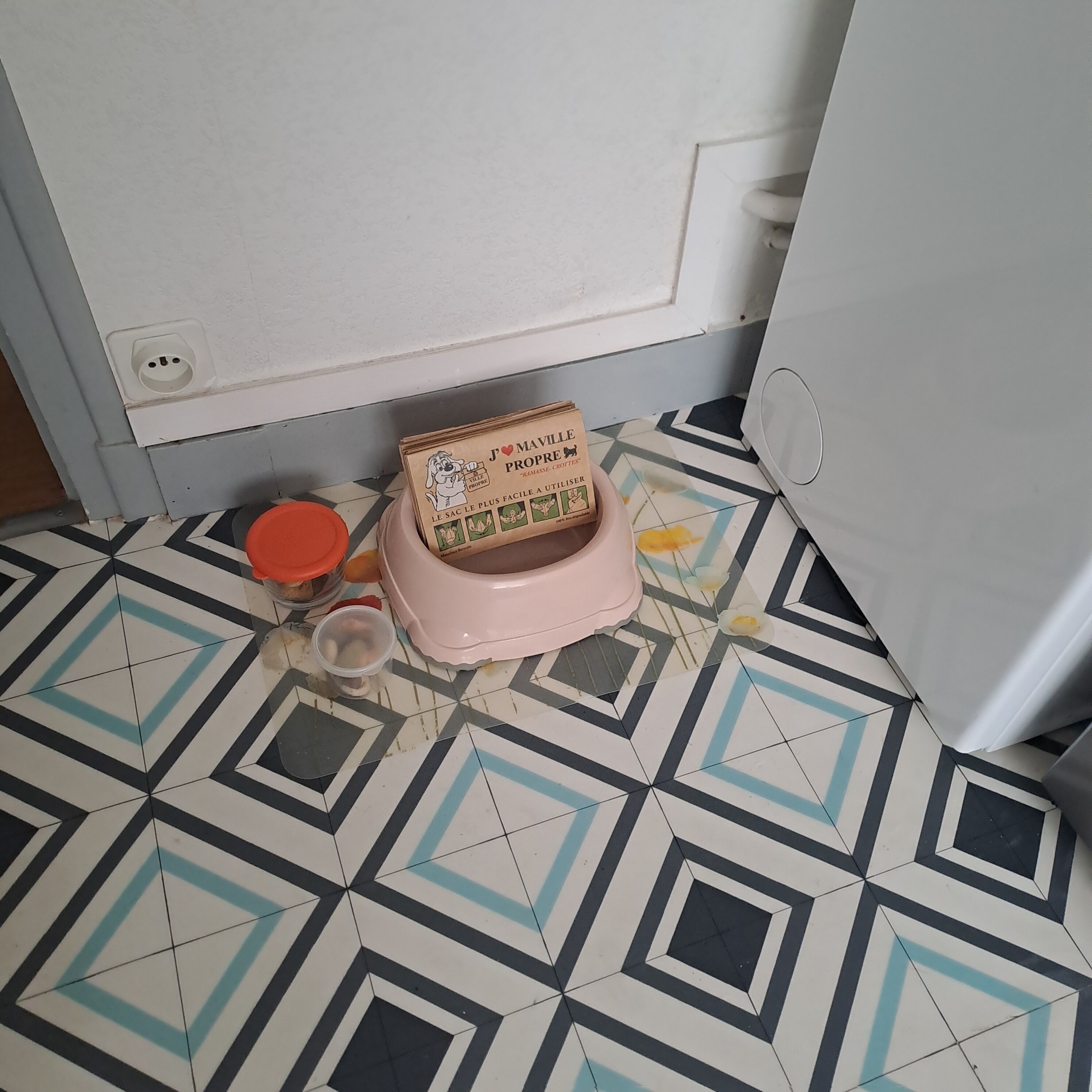 Een keuken met een patroon vloer, een roze houder, een pot met oranje deksel, en een wit apparaat.