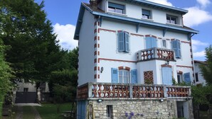 Un bâtiment à deux étages, avec une base en pierre et des volets bleus, entouré d’un jardin bien entretenu où poussent des fleurs violettes.