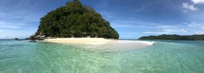 A beach with clear turquoise water, a small sandy island with a gazebo, and lush green vegetation.