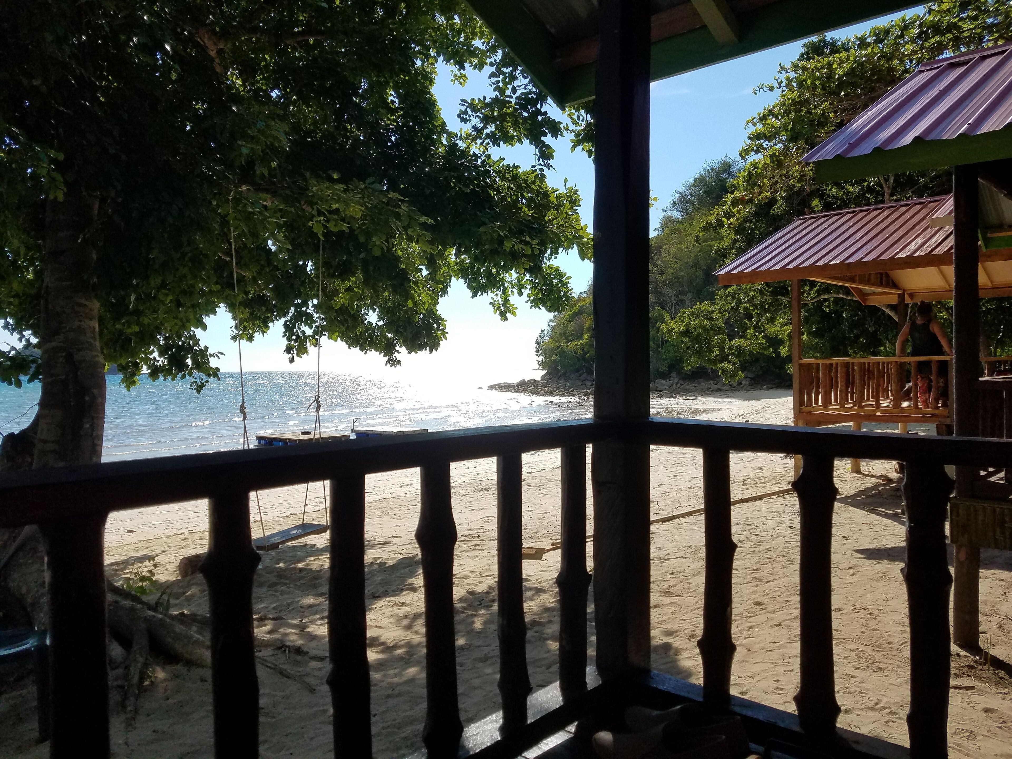 A beach view from a wooden deck with a metal railing, overlooking a sandy beach and the ocean.