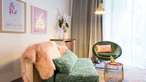 A cozy room with a green upholstered sofa, a fluffy pink throw, a green chair, and a small side table with a vase of flowers. - Nikolai Homes (Vienna)