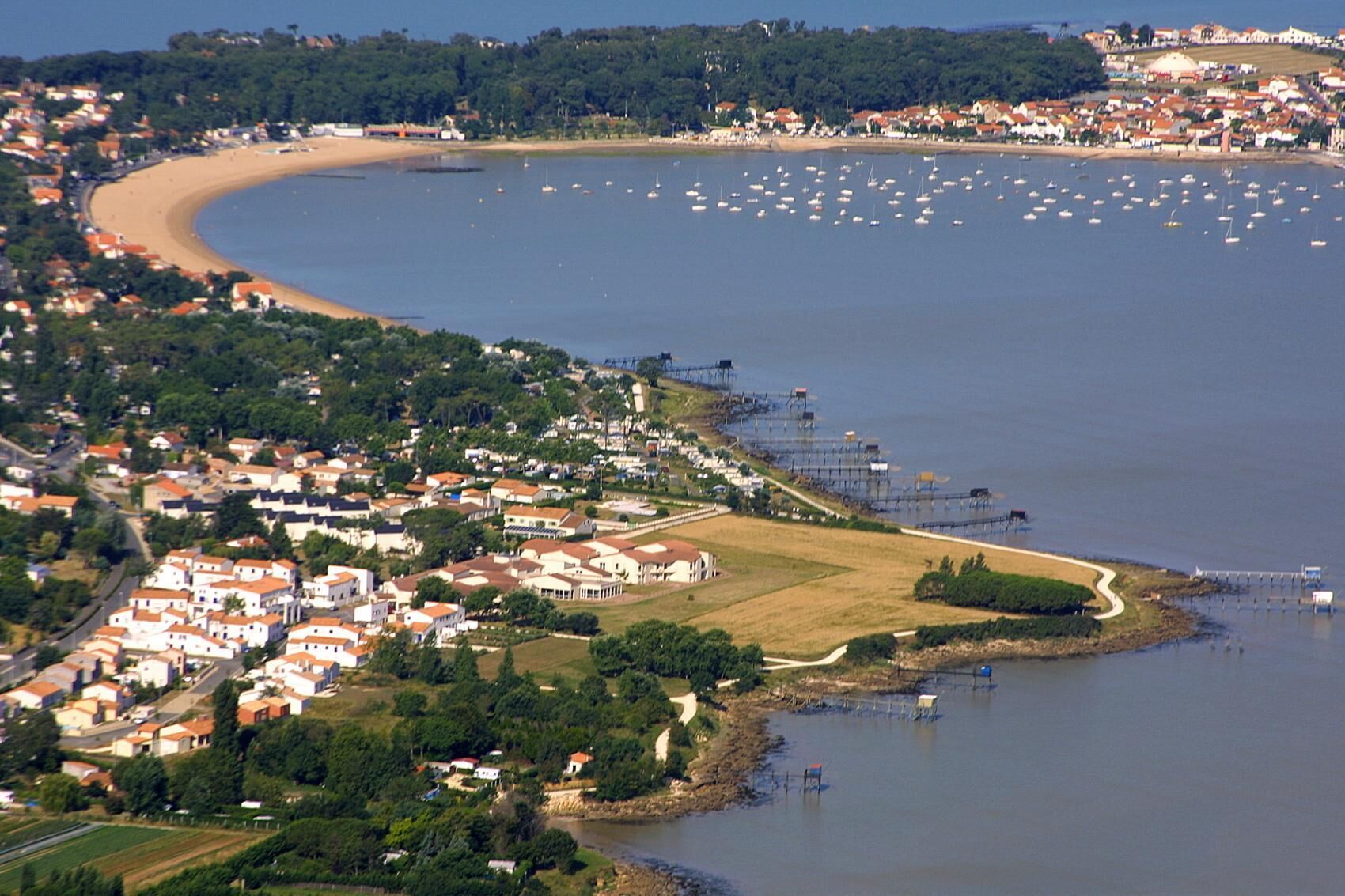 Vue aérienne d’une ville côtière avec une plage, des zones résidentielles et des bateaux sur l’eau.