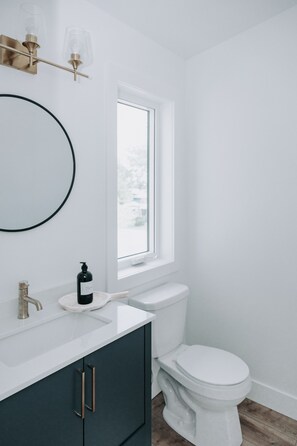 A modern bathroom with a white sink, dark cabinets, a white toilet, and a window above the toilet. - 180 degree lake view, luxury Port Stanley escape overlooking the beach! (Port Stanley)