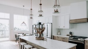 A modern kitchen with a large island, bar stools, hanging pendant lights, and a view of the outdoors through large windows.