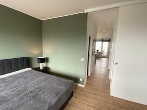 A bedroom with a bed, a nightstand, and a lamp. There is a hallway leading to another room.
