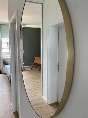 Un pasillo con un espejo grande que refleja una habitación con una cama, un sillón y una puerta.