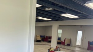 Een ruime kamer met een hoge plafond, een zichtbare vluchtdeur en een zithoek met een bank en stoelen.