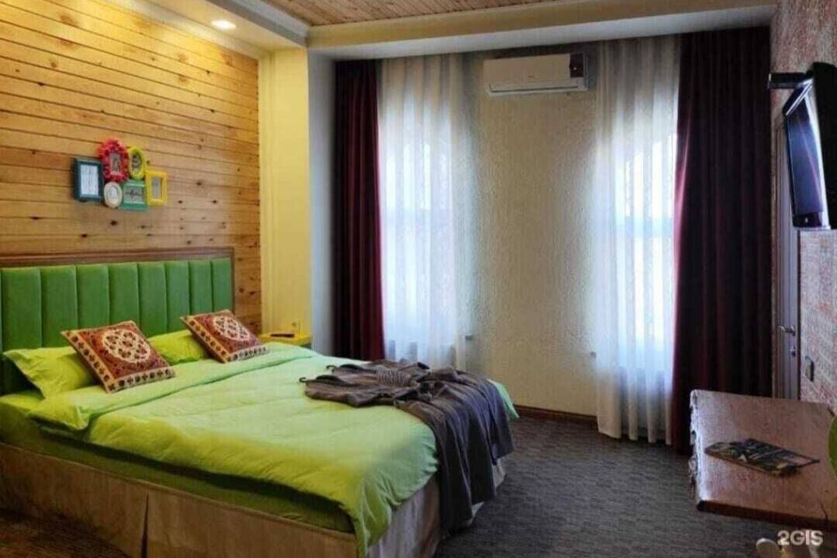 A hotel room with a wooden headboard, a bed with green bedding, a small table with a book, and a window with curtains.