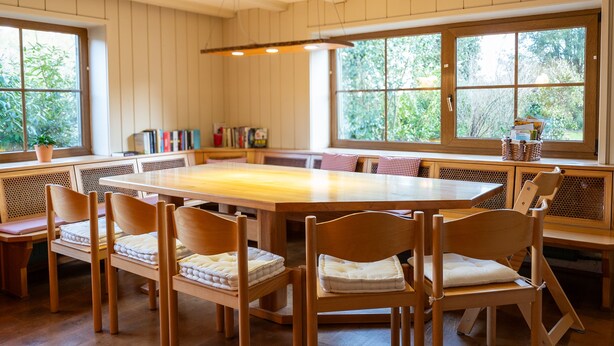 Un comedor con una gran mesa de madera rodeada de sillas, una estantería y un ventanal con vistas a la vegetación.