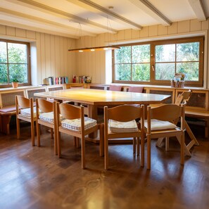 A dining area with a large wooden table surrounded by chairs, a bookshelf, and a window with a view of greenery.