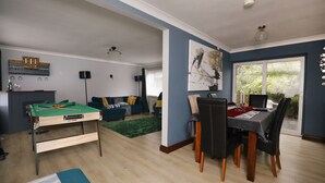 Een woonkamer met een pooltafel, een eethoek met een tafel en stoelen, een zithoek en een groot raam met uitzicht op groen.