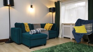 Een woonkamer met een smaragdgroene bank, een klein voetenbankje en een stoel met een blauwe bekleding. Twee vloerlampen, een groen vloerkleed en een raam met gordijnen.