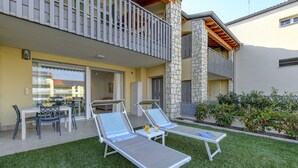 A modern outdoor seating area with two lounge chairs and a table, a stone pillar, and a balcony with a wooden pergola.
