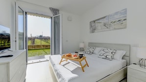 A bedroom with a bed, a bedside table, a TV, and a balcony with a view of the outdoors.