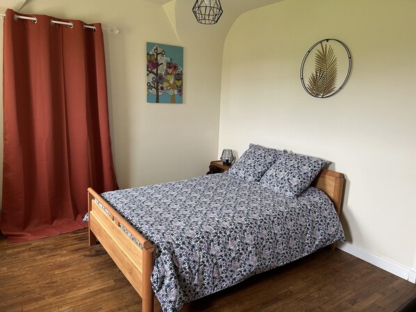 Un dormitorio con una cama de madera, una cortina roja, un cuadro de pájaros, un adorno circular en la pared y una mesita de noche con una lámpara.