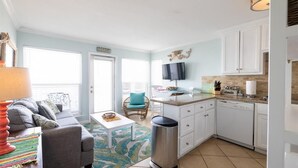 Un espacio habitable compacto con kitchenette, un sofá gris, una pequeña mesa de centro y un cubo de basura.