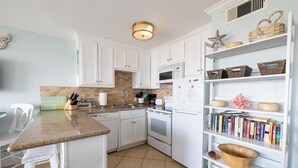 Una cocina con armarios blancos, encimera de granito, microondas y estantería con libros y objetos decorativos.