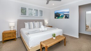 Een slaapkamer met een bed, nachtkastjes, een plafondventilator, een raam met jaloezieën, een aan de muur bevestigd schilderij en een vloerbedekking.