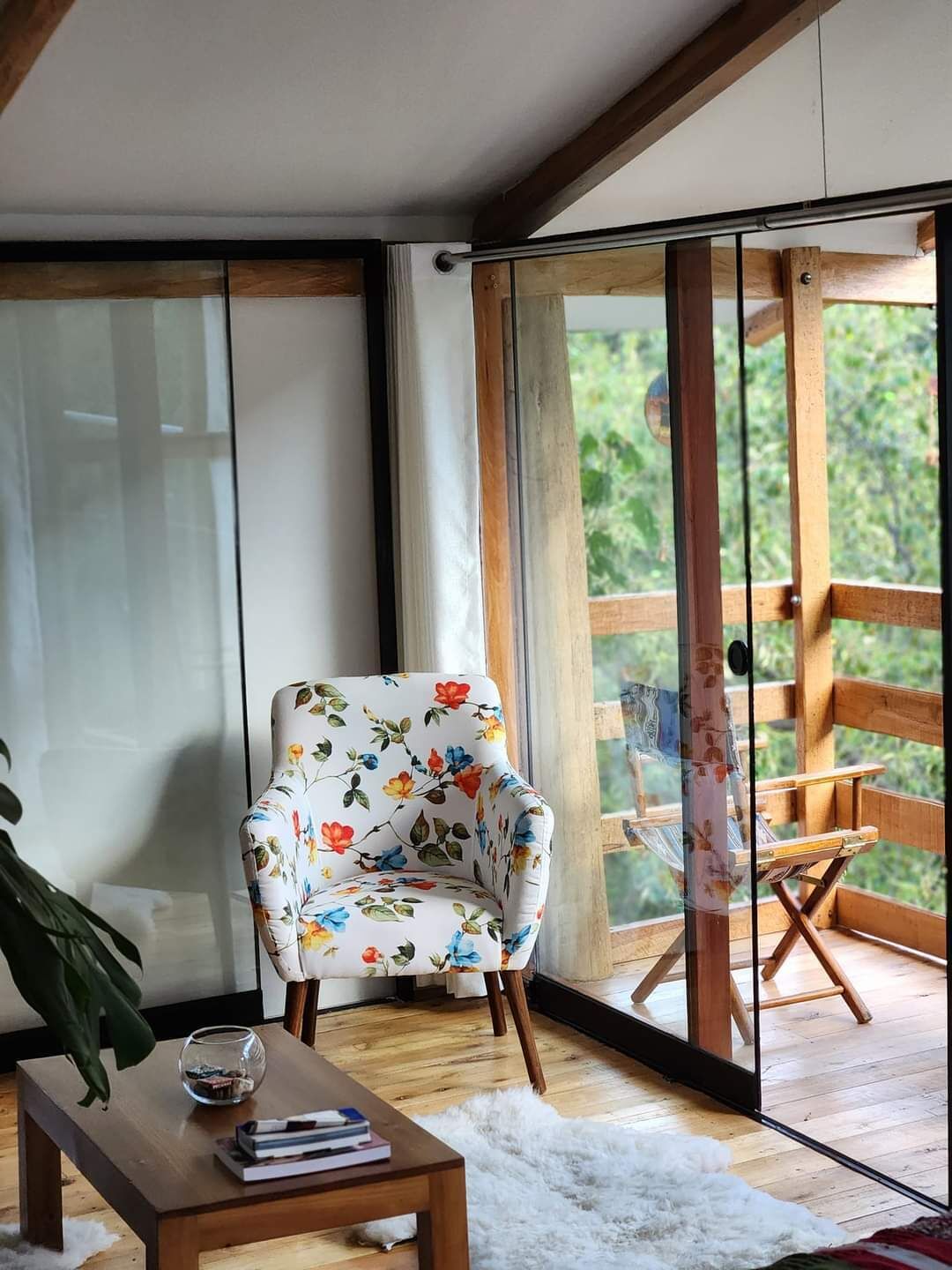 Una habitación con piso de madera, un sillón blanco con estampado floral, una mesa de vidrio con libros y vistas al verdor a través de amplios ventanales.