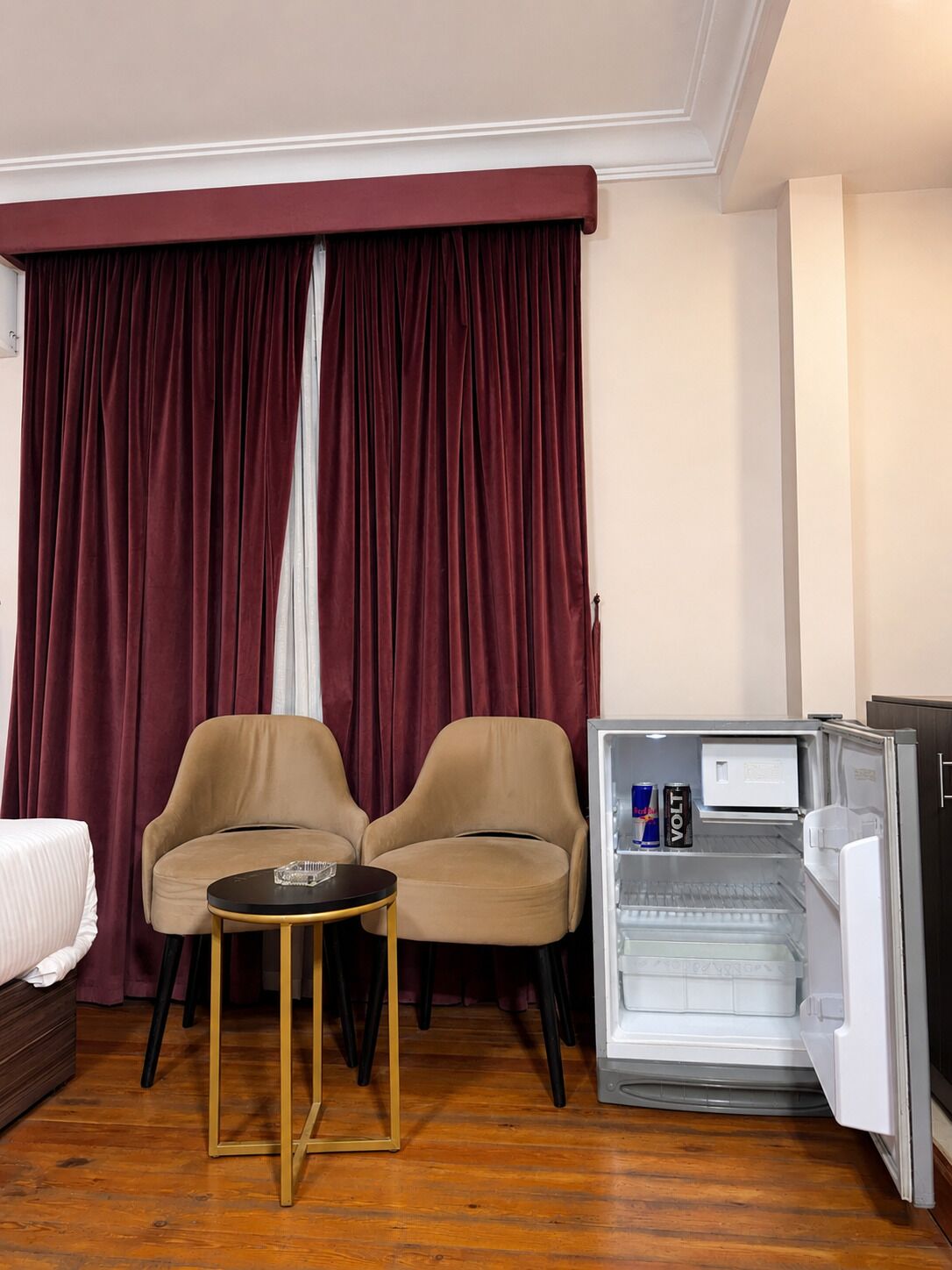 A hotel room with a bed, two armchairs, a small table, a refrigerator, and a window with curtains.