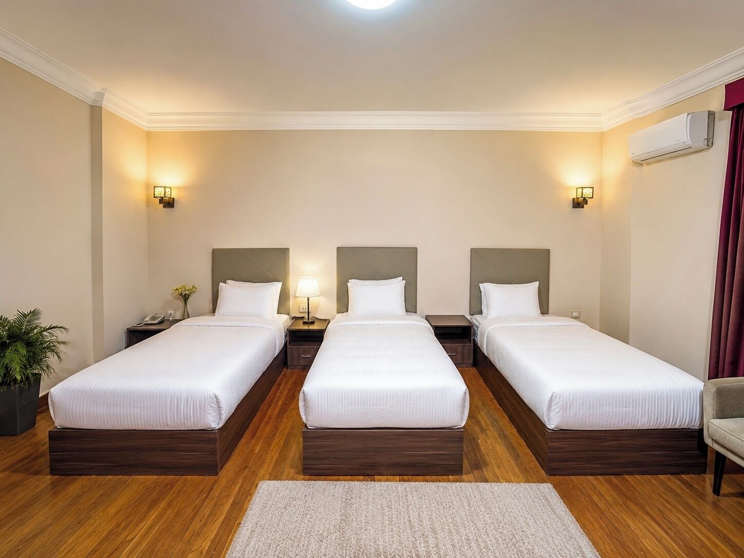 Une chambre d’hôtel avec trois lits simples, un plancher en bois, une petite plante et un bureau avec une lampe.