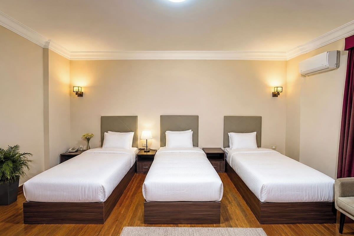 Habitación de hotel con tres camas individuales, suelo de madera, una planta pequeña y un escritorio con lámpara.