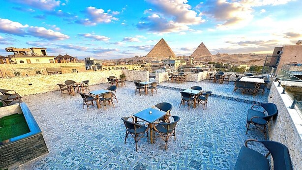 A rooftop terrace with tables and chairs, a patterned floor, and views of pyramids. - Aziz Egypt Pyramids Hotel (Giza)