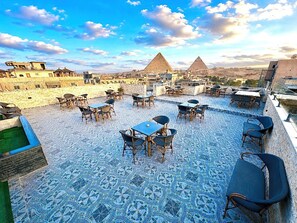 A rooftop terrace with tables and chairs, a patterned floor, and views of pyramids. - Aziz Egypt Pyramids Hotel (Giza)