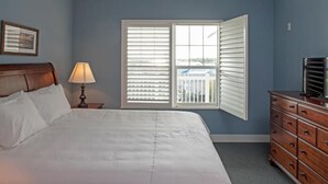 Un dormitorio con cama, mesita de noche con lámpara, cómoda y ventana con persianas.