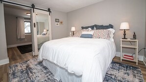 A bedroom with a large bed, a bedside table with a lamp, a mirror, and a door leading to another room. - Charming 3-bedroom, 2 bathroom house with outdoor space in Winona (Winona)