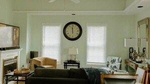 Een woonkamer met een open haard, een bank, een bijzettafeltje, een televisie en een klok aan de muur.