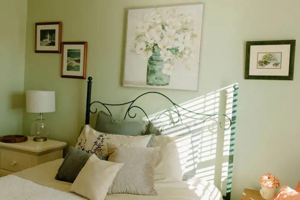 Ein Schlafzimmer mit einem Bett, Kissen, einem Nachttisch, einer Lampe und gerahmten Bildern an der Wand.