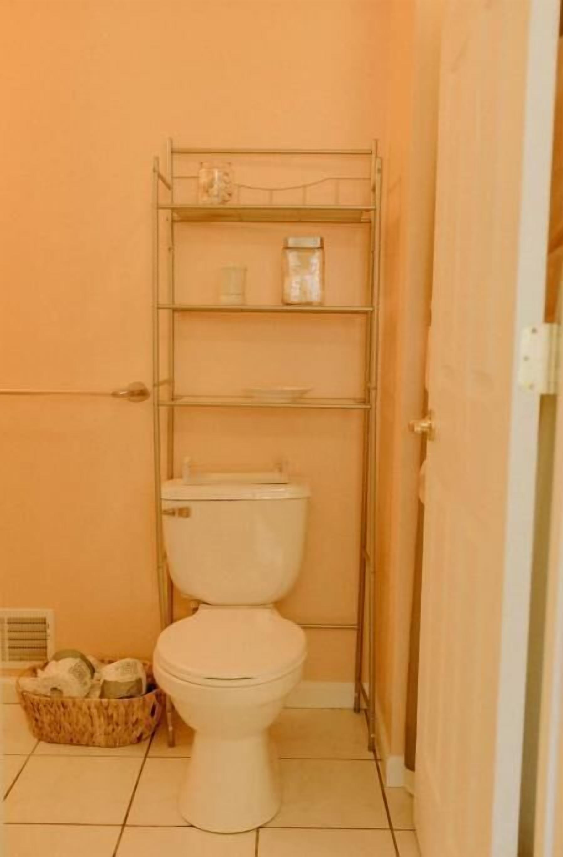 A small bathroom with a toilet, a metal shelf unit above the toilet, and a wicker basket on the floor.