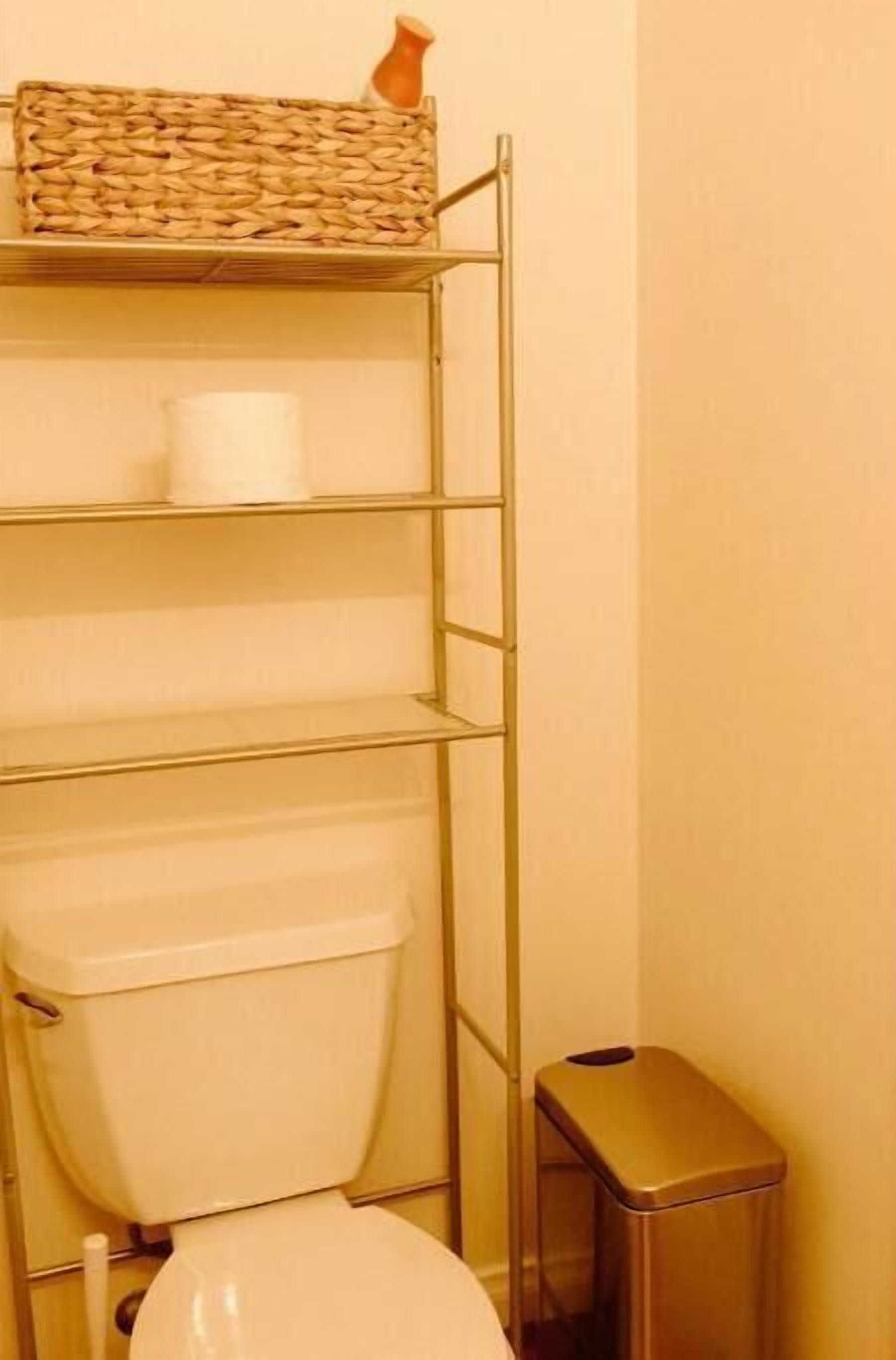 A bathroom with a toilet, a metal shelf unit with a basket and toilet paper, and a trash bin.