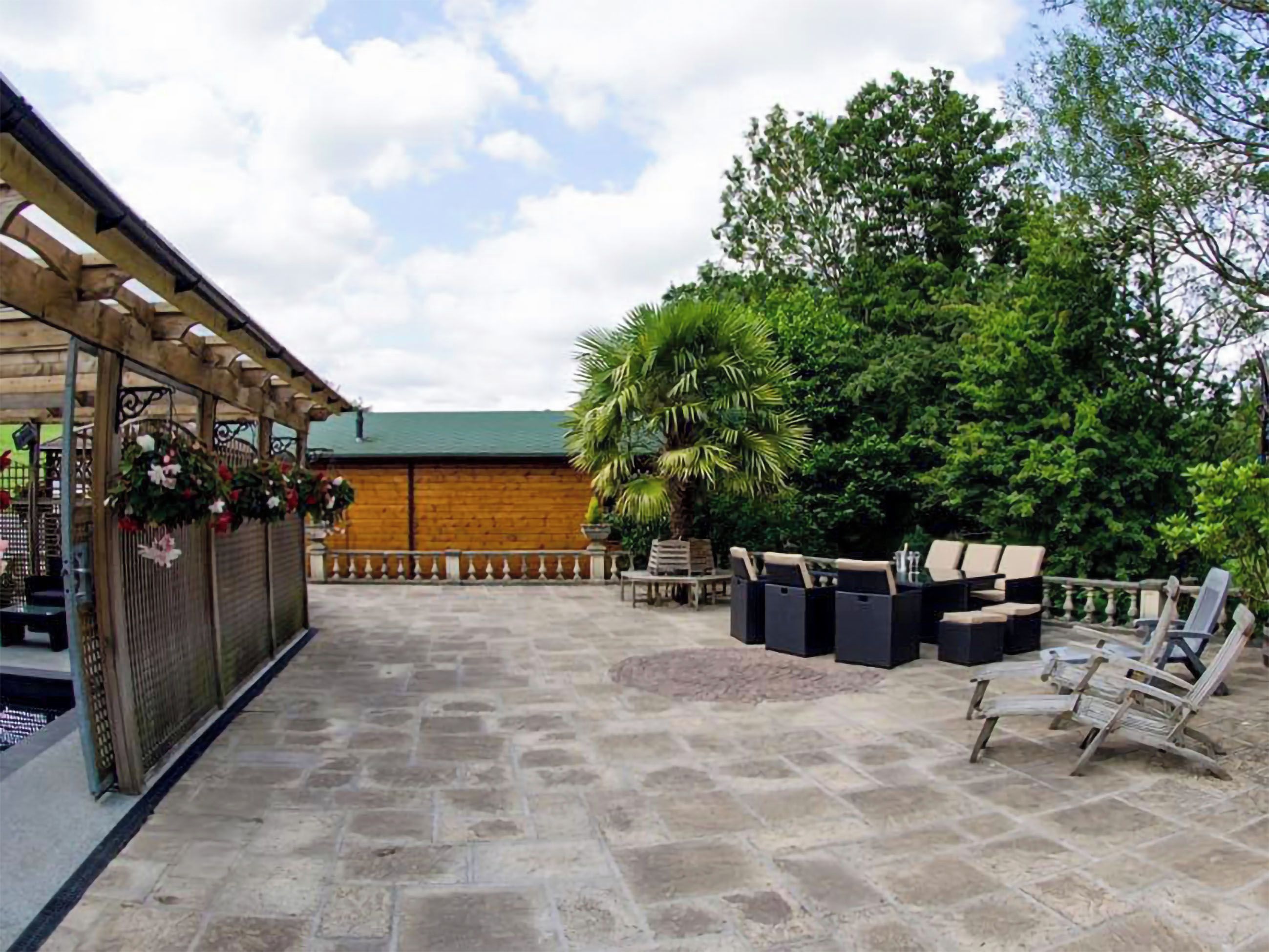 A patio area with stone flooring, outdoor seating, and a wooden pergola with hanging flowers.