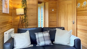A wooden cabin interior with a black leather sofa, a floor lamp, and a framed picture on the wall. - 1 bedroom accommodation in Camerton, near Bath (Camerton, near Bath)