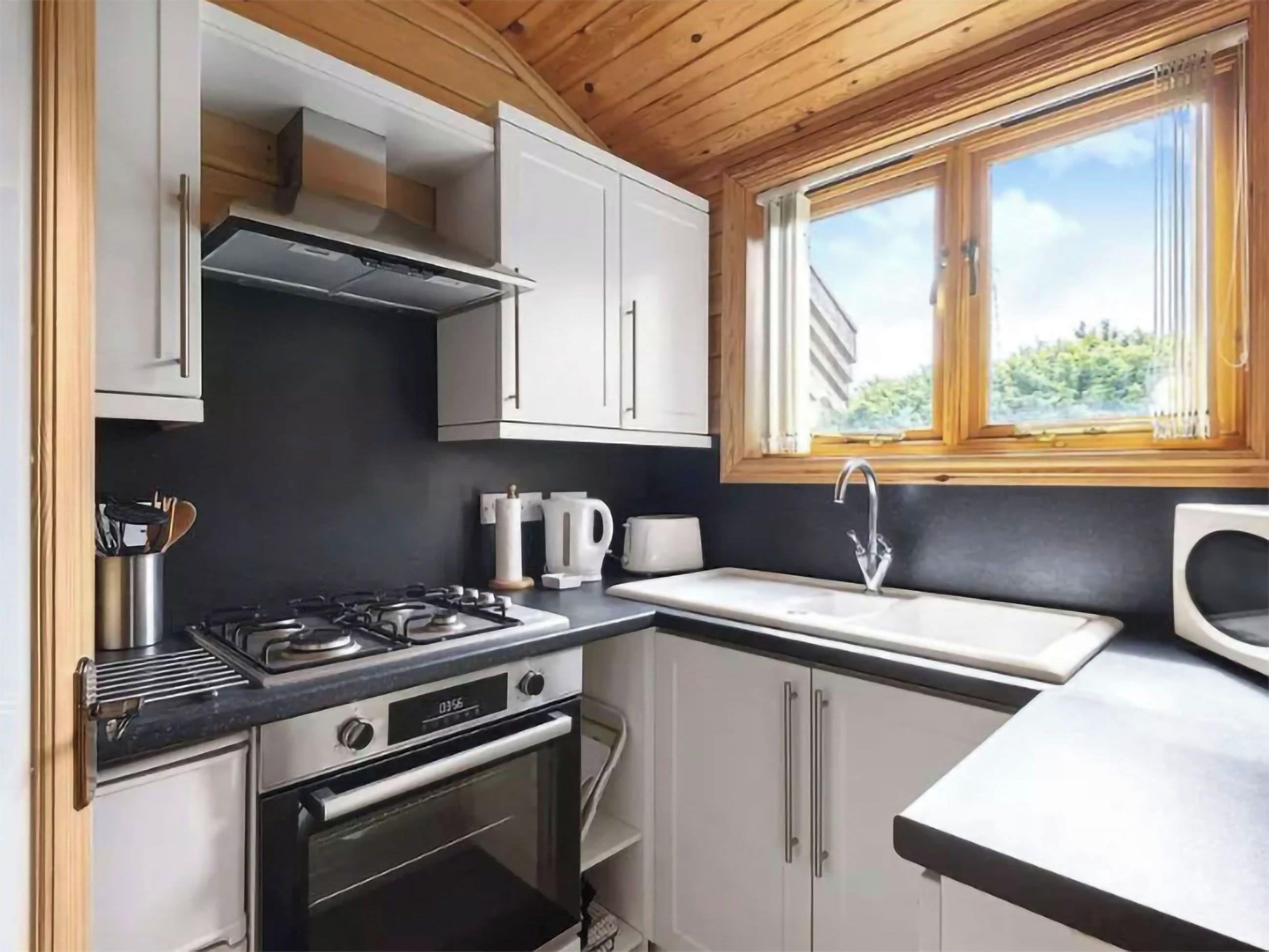 Een kleine keuken met een gasfornuis, oven, witte kastjes en een raam met uitzicht op groen.