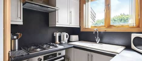 Cocina compacta con estufa a gas, horno, armarios blancos y ventana con vistas al verdor.